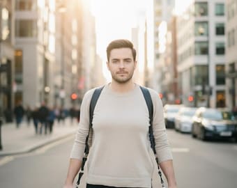 May include: A photographer stands in a city street, holding two cameras. He wears a light beige sweater and black pants. The cameras are black, and he has a black camera strap. The background includes buildings and sunlight.