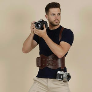 May include: A brown leather camera belt with two buckles and a small pouch. A black camera hangs from the belt, and another camera is held in the hands. The person is wearing a navy blue shirt and khaki pants.