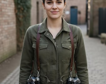 May include: A woman wearing a dark green jacket with a brown leather camera strap holding two black and silver film cameras. The cameras are hanging at chest level. The background is a brick wall and a cobblestone path.