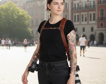 May include: A brown leather camera harness with two cameras attached. The harness is worn over a black t-shirt and black jeans. The background is a city square with people and buildings.