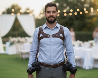 Può includere: Imbracatura per fotocamera in pelle marrone con due fotocamere attaccate. L'imbracatura ha un design per petto e spalle con cinghie regolabili. Il fotografo indossa una camicia azzurra e pantaloni grigi. Lo sfondo mostra un ricevimento di nozze.