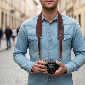 May include: A brown leather camera strap is draped around the neck of a person wearing a light blue denim shirt. The strap supports a black and silver film camera. The background shows a cobblestone street and blurred figures.