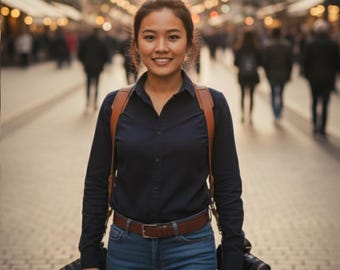 May include: A person wearing a navy blue button-down shirt, blue jeans, and a brown leather camera strap. Two cameras are attached to the strap. The person is smiling and standing on a crowded street.
