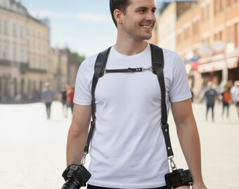 May include: A black leather dual camera harness with two cameras attached. The harness is worn over the shoulders and across the chest. The cameras are black with silver accents. The person is wearing a white t-shirt and black pants.