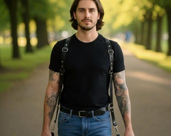May include: A black leather camera harness with two cameras. The harness has adjustable straps and metal buckles. The person is wearing a black t-shirt and blue jeans. The background is a tree-lined path.