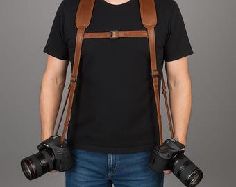 May include: A brown leather dual camera harness with adjustable straps and metal hardware. The harness is designed to hold two cameras, visible with black lenses, and is worn over the shoulders. The photographer is wearing a black t-shirt and blue jeans.