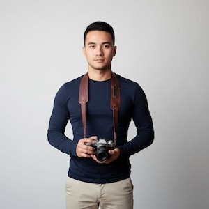 May include: A person wearing a navy blue long-sleeve shirt and khaki pants, holding a silver camera with a brown leather strap around their neck. The camera has a black lens.