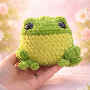 May include: A handmade crochet frog plushie in shades of green and yellow. The frog has large black eyes and small feet. The plushie is held in a hand against a soft, blurred background with flowers, suggesting a whimsical, nature-inspired theme.