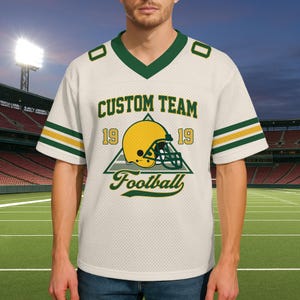 May include: Cream-colored football jersey with green and gold accents. The jersey features the text "CUSTOM TEAM" above a graphic of a football helmet and the word "Football". The jersey has a V-neck and short sleeves.