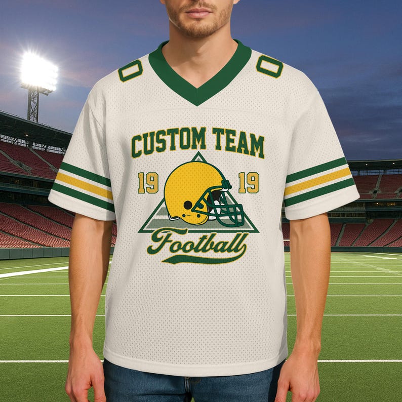 May include: Cream-colored football jersey with green and gold accents. The jersey features the text "CUSTOM TEAM" above a graphic of a football helmet and the word "Football". The jersey has a V-neck and short sleeves.