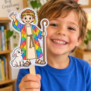 May include: A paper puppet on a wooden stick featuring a cartoon shepherd with curly brown hair, wearing a colorful diamond-patterned robe and a beige tunic, holding a brown staff, and standing next to a small white lamb with pink ears.