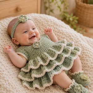 May include: A sage green crochet baby dress featuring three tiered ruffled layers with cream-colored trim, a matching headband with a central flower, and two crochet booties, each adorned with a small flower and a central white pearl accent.