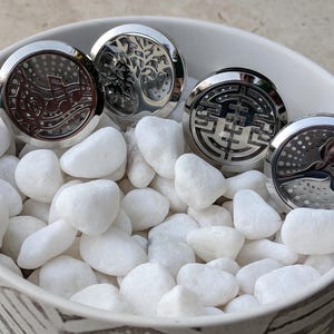 May include: Four silver aromatherapy diffuser lockets with various designs: musical notes, a tree, a geometric pattern, and a bird. The lockets are in a white bowl filled with white stones.