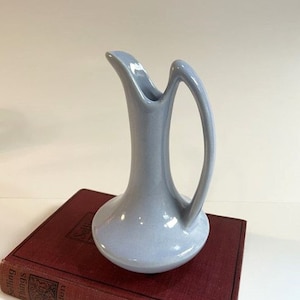 May include: A light blue ceramic pitcher with a wide base and a curved handle. The pitcher is sitting on a red book.