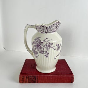 May include: Cream-colored pitcher with a purple floral design. The pitcher has a handle and a decorative rim with a floral pattern. The pitcher is sitting on a red book.