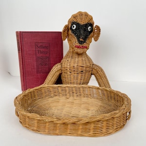 May include: A woven wicker monkey figure with a black face mask, white eyes, and red lips, positioned behind an oval-shaped woven wicker basket. Beside the monkey stands a red hardcover book with the text Selling Things on the front cover.