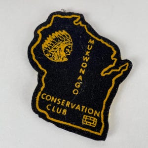 May include: Black felt patch in the shape of the state of Wisconsin, with a gold border. The patch features a gold Native American profile, the words "Mukwonago Conservation Club," and a small graphic.