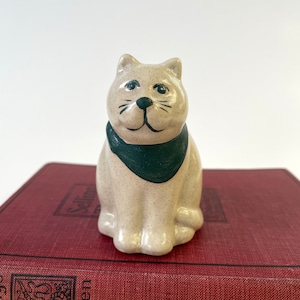 May include: A small ceramic cat figurine with a green bandana. The cat is sitting on a red book with black text.