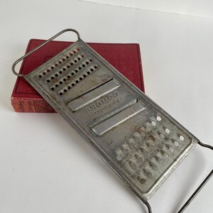 May include: A vintage metal cheese grater with the brand name "BROMCO" stamped on the surface. The grater has a rectangular shape with multiple small holes and a handle. It rests on a red book.
