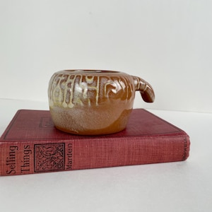 May include: A small, brown ceramic mug with a handle. The mug has a rounded shape and a textured, glazed surface with lighter-colored accents. It sits atop a red book with the title "Selling Things" visible.