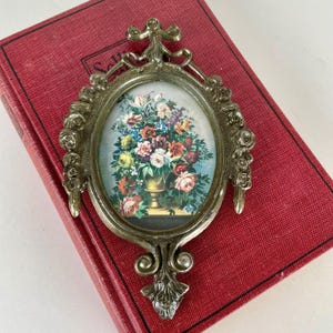 May include: An oval, silver-toned picture frame with a floral still life. The frame has ornate detailing and is set against a red book. The artwork features a vase overflowing with colorful flowers, including roses and other blooms.