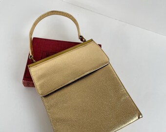 May include: A gold-colored, rectangular handbag with a matching handle. The bag has a flap closure and a textured surface. The handle is arched and attached to the top of the bag with gold-toned hardware. The bag is resting on a red book.