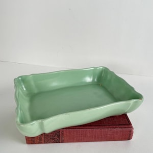 May include: A rectangular, light green ceramic dish with a scalloped edge. The dish is resting on a red book. The dish has a glossy finish and a slightly curved shape. The overall aesthetic is vintage.