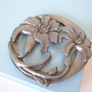 May include: A silver metal coaster with a design of two lilies. The lilies are in a circular design with the stems intertwined.