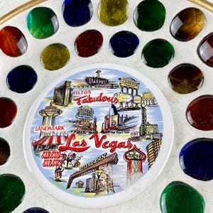 May include: A decorative plate featuring a Las Vegas themed design with iconic landmarks and the text "Las Vegas". The plate is surrounded by colorful glass stones in various colors, including red, green, and blue. The word "Fabulous" is also visible.