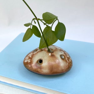 May include: A small, brown and white ceramic vase with multiple holes for stems. The vase is shaped like a rounded rock and is sitting on a blue book.