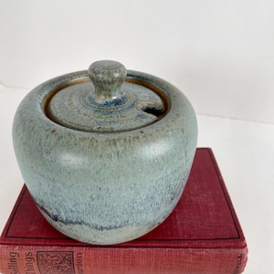 May include: A round ceramic sugar bowl with a lid featuring a small knob handle and a side notch for a spoon. The glaze is a mottled blend of sage green, dusty blue, and earthy brown tones, creating a textured, variegated appearance on the surface.