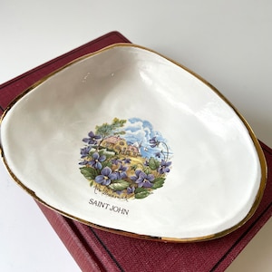 May include: White ceramic dish with a gold rim, featuring a colorful illustration of a coastal scene with buildings, flowers, and the text "SAINT JOHN" and "New Brunswick". The dish is placed on a burgundy surface.