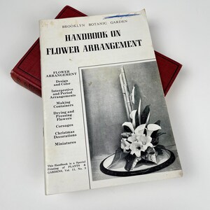 May include: A black and white photo of a book titled "Handbook on Flower Arrangement" published by Brooklyn Botanic Garden. The book cover features a floral arrangement with white flowers, green leaves, and bamboo stalks.
