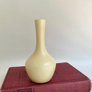 May include: A cream-colored, slender vase with a rounded base and a long, narrow neck. The vase is made of a smooth material and sits atop a vintage, red book with gold lettering.