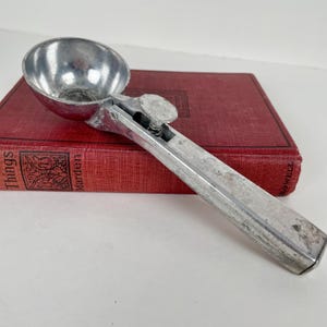 May include: A silver-colored metal ice cream scoop with a rounded bowl and a spring-loaded release mechanism on the handle. The scoop features a textured, weathered finish and is positioned diagonally across a rectangular red hardcover book.