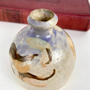 May include: A small, round ceramic vase with a narrow neck. The vase is painted with a light blue background and brown and tan abstract designs. The vase is a unique piece of pottery.