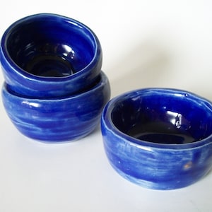 May include: Three small blue ceramic bowls with a white glaze. The bowls are stacked on top of each other, with the top bowl slightly tilted to the side. The bowls have a slightly uneven, handmade look.