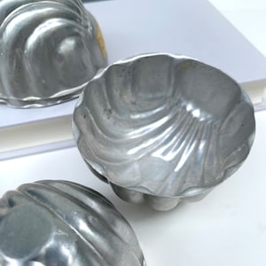 May include: Three vintage, silver-colored metal bundt cake molds. Each mold has a fluted, scalloped design. The molds are arranged on a white surface, with a book visible in the background. The molds are likely made of aluminum.