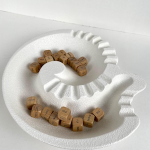 May include: A white, textured, ceramic dish in a spiral design, filled with small wooden dice. The dice have various words printed on them. The dish is on a white surface.