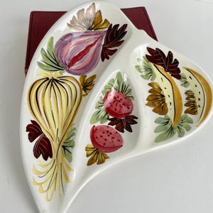May include: A white ceramic divided serving dish with a three-section leaf shape. The surface features hand-painted illustrations of a yellow onion, two red tomatoes, and a yellow fruit slice, all accented with dark red and green foliage details.
