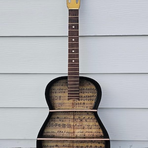 May include: A decorative guitar-shaped shelf with a black frame and a gold-colored headstock. The interior features sheet music, and it has two shelves. The bottom of the shelf has a gold decorative design.