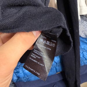 May include: Close-up of a navy blue knit sweater with a tag. The tag reads "ALLSAINTS.COM" and includes the text "FRITZ HOODY" and "MK086C". The tag also indicates the sweater is made of 100% cotton.