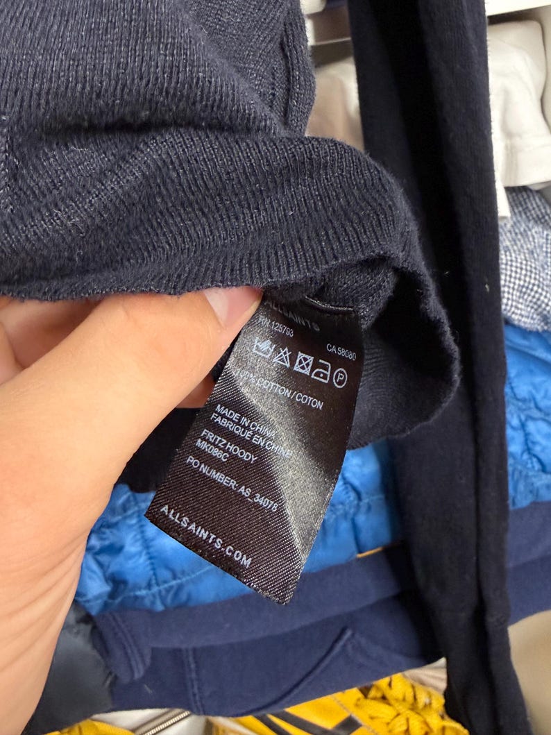 May include: Close-up of a navy blue knit sweater with a tag. The tag reads "ALLSAINTS.COM" and includes the text "FRITZ HOODY" and "MK086C". The tag also indicates the sweater is made of 100% cotton.