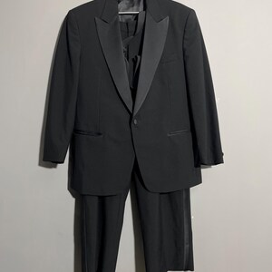 May include: A formal black tuxedo suit, including a jacket with satin lapels, a vest, and matching trousers. The suit is hanging on a hanger against a neutral background. The trousers have a satin stripe down the side.