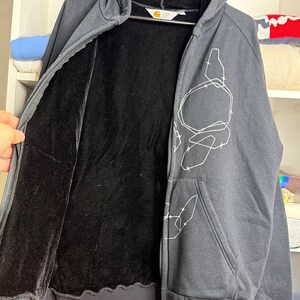 Pode incluir: Casaco com capuz e z&iacute;per cinza carv&atilde;o com forro interno preto e design gr&aacute;fico branco. O casaco tem capuz, bolso frontal e bainha e punhos canelados pretos. O logotipo da Carhartt &eacute; vis&iacute;vel.