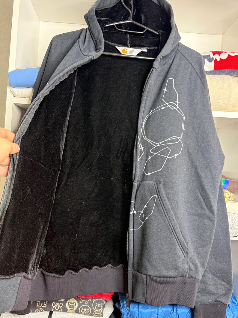 Pode incluir: Casaco com capuz e z&iacute;per cinza carv&atilde;o com forro interno preto e design gr&aacute;fico branco. O casaco tem capuz, bolso frontal e bainha e punhos canelados pretos. O logotipo da Carhartt &eacute; vis&iacute;vel.