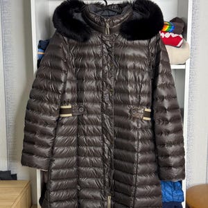 May include: A brown puffer coat with a black fur-trimmed hood. The coat has a quilted design, a zipper closure, and decorative accents. The coat is hanging on a black hanger.