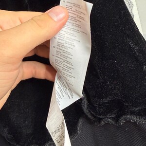 Pode incluir: Close-up de uma pe&ccedil;a de roupa preta com textura aveludada. Uma etiqueta branca com texto em v&aacute;rios idiomas &eacute; vis&iacute;vel, detalhando a composi&ccedil;&atilde;o do tecido e as instru&ccedil;&otilde;es de cuidado. A pe&ccedil;a de roupa parece ser um casaco ou um sobretudo.