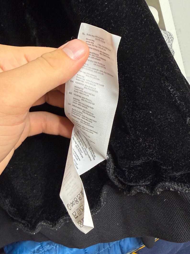 Pode incluir: Close-up de uma pe&ccedil;a de roupa preta com textura aveludada. Uma etiqueta branca com texto em v&aacute;rios idiomas &eacute; vis&iacute;vel, detalhando a composi&ccedil;&atilde;o do tecido e as instru&ccedil;&otilde;es de cuidado. A pe&ccedil;a de roupa parece ser um casaco ou um sobretudo.