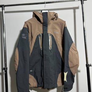 Könnte beinhalten: Braune und schwarze Kapuzenjacke mit Reißverschluss. Die Jacke hat einen hohen Kragen und mehrere Taschen. Das Bergans-Logo ist auf der Brust sichtbar. Die Jacke hängt an einem Metallgestell.